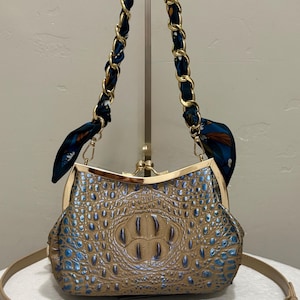 May include: A tan and blue crocodile-embossed handbag with a gold-tone metal kiss-lock frame closure. The bag features a gold chain strap interwoven with a dark blue patterned fabric scarf and an additional adjustable tan leather shoulder strap.