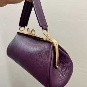 May include: A deep purple handbag with a textured surface, featuring a gold-toned metal frame closure with a small bow-shaped clasp. The bag has a single matching purple handle attached to the frame with gold-toned rings, creating a structured shape.