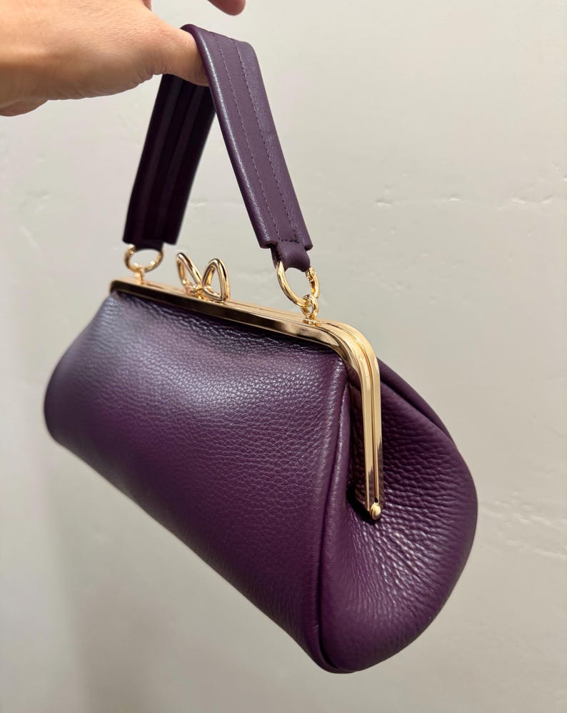 May include: A deep purple handbag with a textured surface, featuring a gold-toned metal frame closure with a small bow-shaped clasp. The bag has a single matching purple handle attached to the frame with gold-toned rings, creating a structured shape.