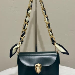May include: A dark green handbag with a gold-tone clasp and chain strap. The strap is adorned with a black and white scarf. The bag has a rectangular shape with a curved flap.