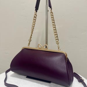 May include: A deep purple handbag with a textured surface, featuring a gold-toned metal frame and a bow-shaped clasp. The bag includes a gold chain link handle and a long, thin purple shoulder strap, providing both handheld and crossbody options.