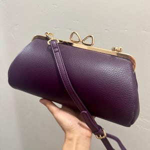 May include: A deep purple textured handbag with a gold-toned metal frame and a bow-shaped clasp. The bag features a matching purple adjustable shoulder strap attached to the sides with gold-toned hardware, creating a structured, elongated shape.