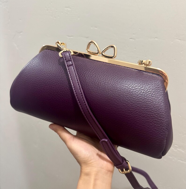 May include: A deep purple textured handbag with a gold-toned metal frame and a bow-shaped clasp. The bag features a matching purple adjustable shoulder strap attached to the sides with gold-toned hardware, creating a structured, elongated shape.