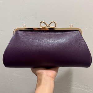 May include: A deep purple clutch bag with a textured surface, featuring a gold-toned metal frame and a bow-shaped clasp. The bag includes two small gold-toned rings on the top frame for attaching a strap, presenting a structured and elegant design.