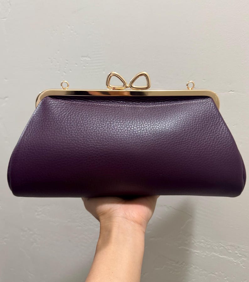 May include: A deep purple clutch bag with a textured surface, featuring a gold-toned metal frame and a bow-shaped clasp. The bag includes two small gold-toned rings on the top frame for attaching a strap, presenting a structured and elegant design.