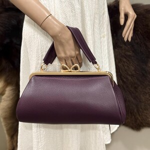 May include: A deep purple handbag with a textured leather-like surface, featuring a gold-toned metal frame and a distinctive bow-shaped clasp. The bag has a single top handle attached with gold-toned hardware, providing a structured silhouette.