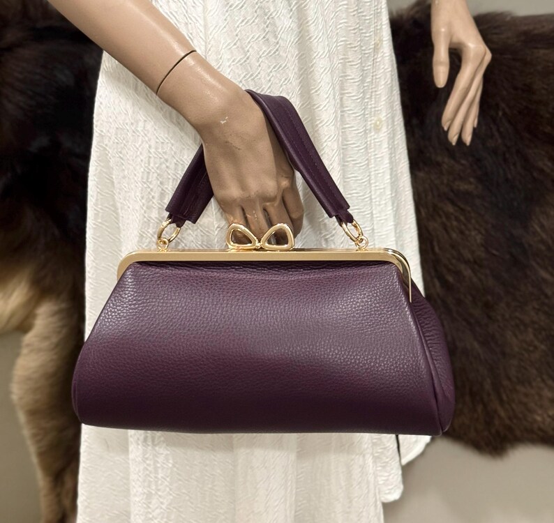 May include: A deep purple handbag with a textured leather-like surface, featuring a gold-toned metal frame and a distinctive bow-shaped clasp. The bag has a single top handle attached with gold-toned hardware, providing a structured silhouette.