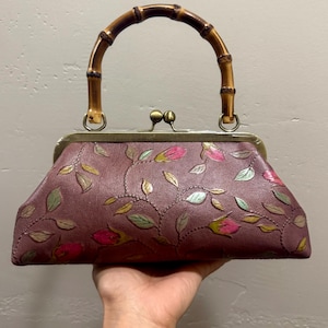 May include: A burgundy leather handbag with a bamboo handle. The bag features an embossed floral design with pink and green accents. The clasp is a gold tone.