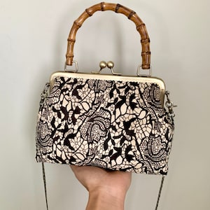 May include: A vintage-style handbag with a bamboo handle and a decorative clasp. The bag features a black and cream-colored lace pattern fabric. It has a metal frame and a detachable chain strap.