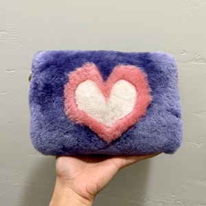 May include: A rectangular, fluffy purple clutch with a heart design. The heart is outlined in pink and filled with white. The clutch is held in a hand, showcasing its size and texture. The bag has a gold zipper.