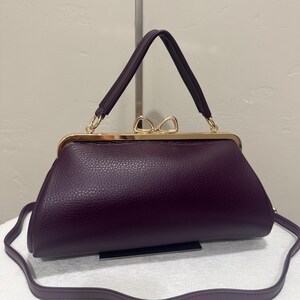 May include: A deep purple handbag with a textured leather-like finish, featuring a gold-toned metal frame closure topped with a decorative bow-shaped clasp, a single top handle, and a detachable thin shoulder strap for versatile carrying options.