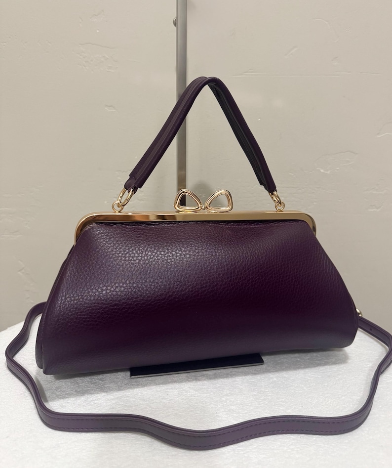 May include: A deep purple handbag with a textured leather-like finish, featuring a gold-toned metal frame closure topped with a decorative bow-shaped clasp, a single top handle, and a detachable thin shoulder strap for versatile carrying options.