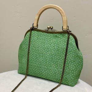 May include: A green fabric handbag featuring a small white floral pattern, a light-colored wooden arch handle, an antique brass-toned metal frame with a kiss-lock clasp, and a detachable thin bronze-colored metal chain shoulder strap.