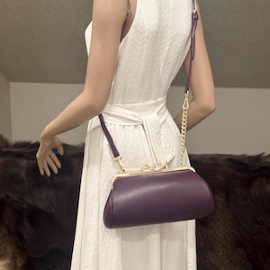 May include: A dark purple, textured, oblong-shaped clutch bag with a gold-toned metal frame, a kiss-lock closure featuring a small bow detail, and a detachable shoulder strap that combines a thin purple strap with a gold-toned metal chain link.