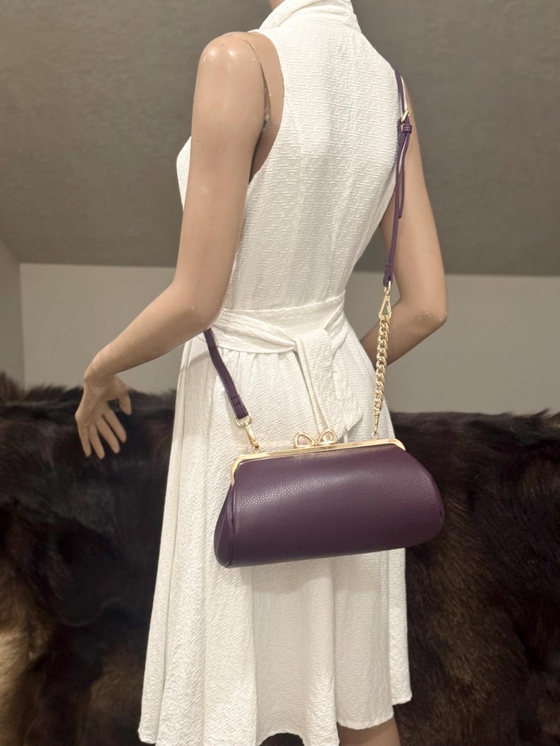 May include: A dark purple, textured, oblong-shaped clutch bag with a gold-toned metal frame, a kiss-lock closure featuring a small bow detail, and a detachable shoulder strap that combines a thin purple strap with a gold-toned metal chain link.