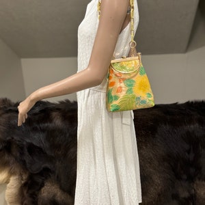 May include: A vintage handbag with a yellow floral design, green leaves, and orange accents. The bag has a gold-colored frame and a yellow chain strap. The bag is held by a mannequin wearing a white dress.