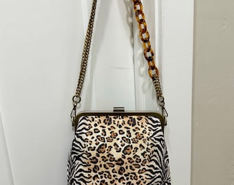 May include: A rectangular handbag with a clasp closure, featuring a mixed animal print design. The bag has a gold-colored chain strap and a tortoiseshell-style handle. The print combines zebra stripes and leopard spots.
