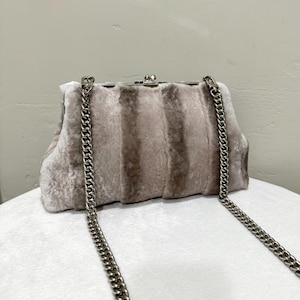 May include: A light gray faux fur clutch with a silver-tone metal clasp and chain strap. The purse has a rectangular shape with vertical stripes and a kiss-lock closure. The chain strap hangs down on both sides.