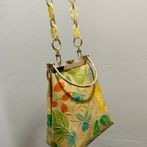 May include: A vintage handbag with a cream-colored background and a floral pattern in green, orange, and blue. The bag has a gold-colored metal frame and handle, with a cream-colored chain strap.