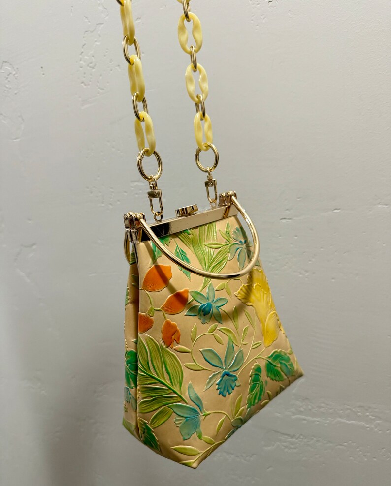 May include: A vintage handbag with a cream-colored background and a floral pattern in green, orange, and blue. The bag has a gold-colored metal frame and handle, with a cream-colored chain strap.