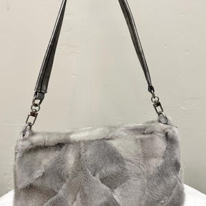 May include: A gray faux fur shoulder bag with a silver chain strap. The bag has a rectangular shape and a textured surface. The strap is made of a silver material. The bag is a fashion accessory.