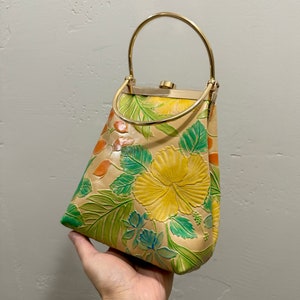 May include: Tan leather handbag with a gold-tone handle and clasp. The bag features embossed floral designs in yellow, green, and orange. The bag is held by a hand.