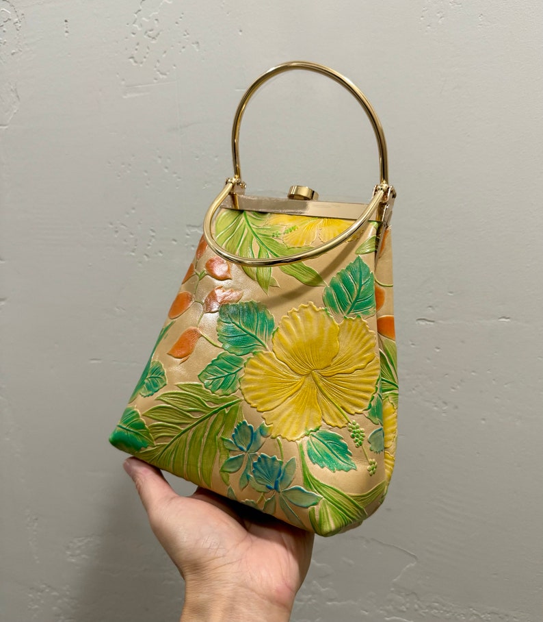 May include: Tan leather handbag with a gold-tone handle and clasp. The bag features embossed floral designs in yellow, green, and orange. The bag is held by a hand.