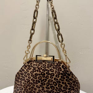 May include: A structured handbag featuring a brown and black leopard print pattern. The bag has a gold-tone metal frame, a top handle, and a detachable chain strap with alternating gold-tone metal and leopard print resin links.