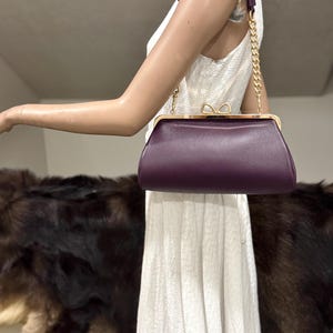 May include: A deep purple textured leather handbag with a gold-toned metal frame and a bow-shaped clasp. The bag features a short handle with a gold-toned chain link section on each side, providing a structured and compact design for carrying.