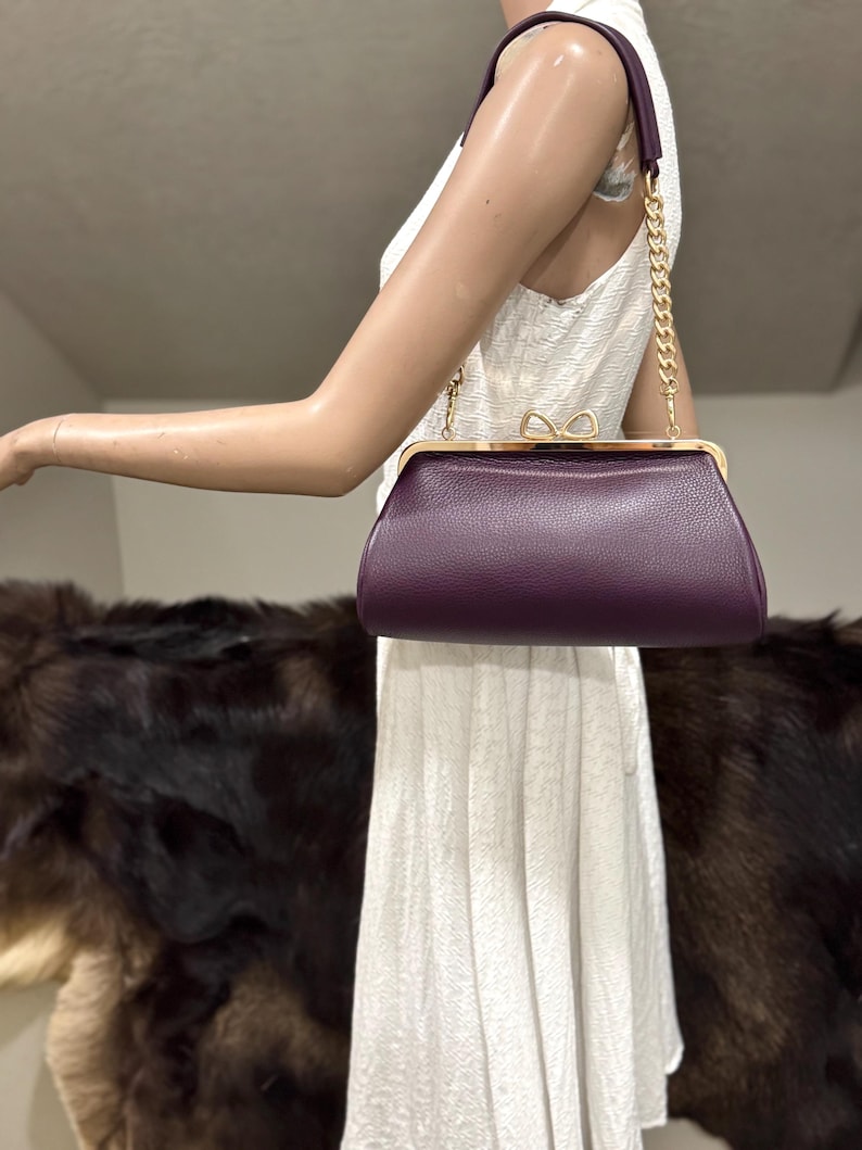 May include: A deep purple textured leather handbag with a gold-toned metal frame and a bow-shaped clasp. The bag features a short handle with a gold-toned chain link section on each side, providing a structured and compact design for carrying.