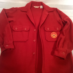 May include: A red wool jacket with a collared neckline, button-down front, and two chest pockets. The jacket features a Boy Scouts of America patch on the right pocket and buttoned cuffs. The jacket is a size 6.