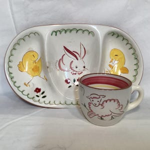 May include: A vintage ceramic Easter-themed serving dish with three sections, featuring a bunny, chick, and duck design. A matching mug with a sheep illustration is also present. The dish has a green border and red accents.