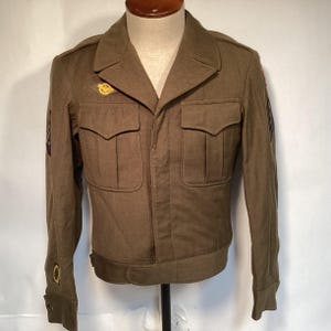 Puede incluir: Chaqueta militar verde oliva con dos bolsillos en el pecho y dos bolsillos inferiores. La chaqueta tiene cuello con solapas, puños abotonados y un emblema dorado en el pecho. Se ven parches en las mangas.