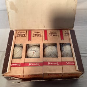 May include: A vintage box of Spalding custom imprinted golf balls. The box is open, revealing four individual brown cardboard containers, each holding golf balls. The containers have red accents and the Spalding logo. The text "Custom Imprinted Golf Balls" is printed on each container.