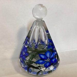 Può includere: Un flacone di profumo in vetro trasparente a forma di goccia. Il flacone ha un tappo trasparente ed è decorato con disegni floreali blu. La base del flacone è più larga della parte superiore e il motivo floreale è visibile attraverso il vetro.