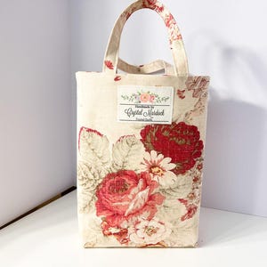 May include: A rectangular tote bag with a floral pattern featuring large red roses and white daisies on a cream background. The bag has matching handles and a label that reads "Handmade by Crystal Murdock".