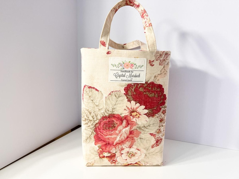 May include: A rectangular tote bag with a floral pattern featuring large red roses and white daisies on a cream background. The bag has matching handles and a label that reads "Handmade by Crystal Murdock".