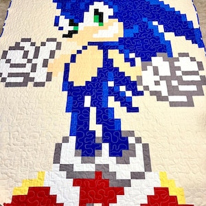 May include: A handmade quilt depicting Sonic the Hedgehog in a pixelated design. The quilt uses blue, white, grey, red, and yellow against a cream backdrop. The quilt is likely made of cotton and has a quilted pattern, suitable for a bed or sofa.