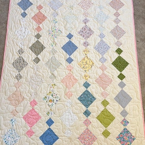 May include: A white quilt with a pink border. The quilt is made up of a series of diamond-shaped patches in various floral patterns, including pink, blue, green, and yellow.