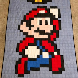 May include: A pixelated quilt featuring a red and blue Super Mario character with a red cap and moustache. The character is standing on a green and black platform with a gold coin above.