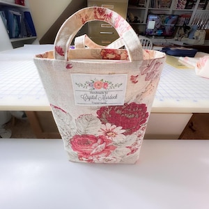 May include: A beige tote bag with a floral pattern of red and white roses. The bag has matching handles and a label that reads "Handmade by Crystal Murdock". The bag is approximately 10 inches tall.