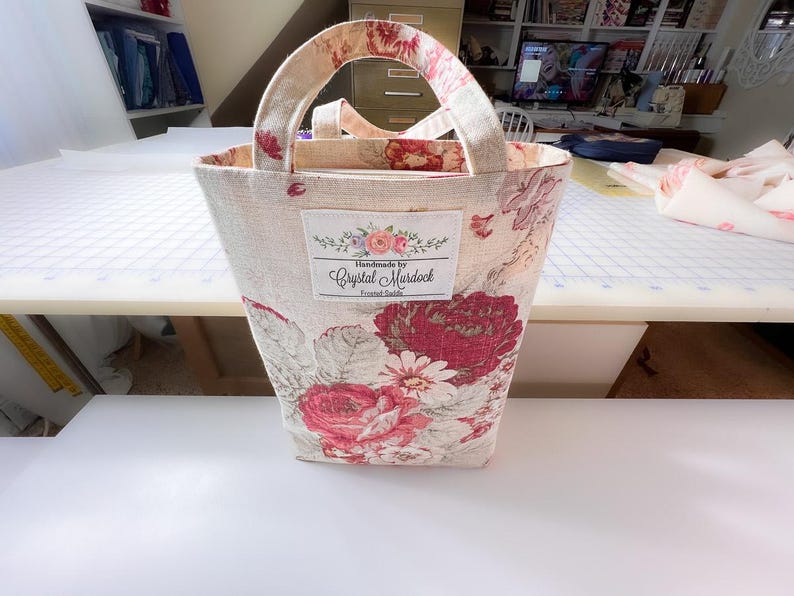 May include: A beige tote bag with a floral pattern of red and white roses. The bag has matching handles and a label that reads "Handmade by Crystal Murdock". The bag is approximately 10 inches tall.
