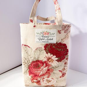 May include: A rectangular tote bag with a floral pattern featuring large red roses and white flowers on a cream background. The bag has a matching handle and a label that reads "Handmade by Crystal Murdock".