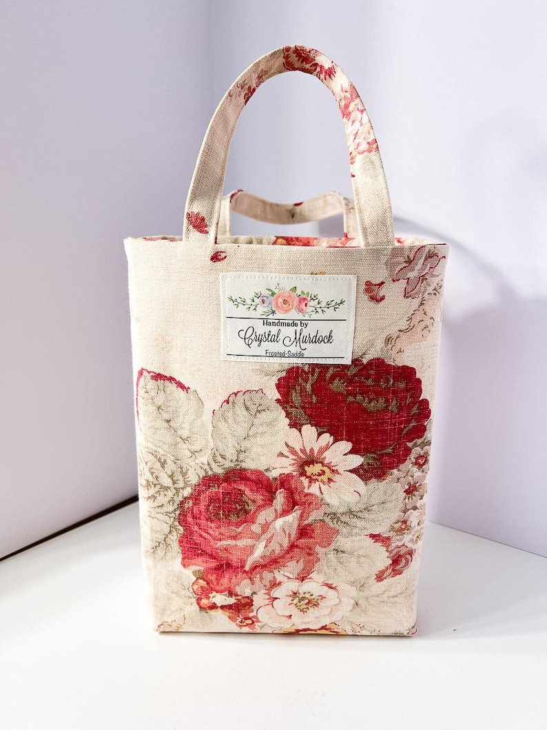 May include: A rectangular tote bag with a floral pattern featuring large red roses and white flowers on a cream background. The bag has a matching handle and a label that reads "Handmade by Crystal Murdock".