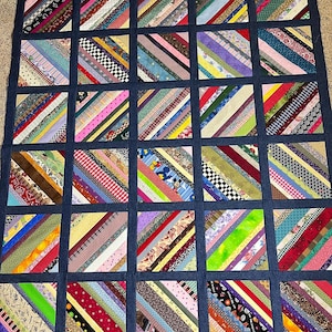 Puede incluir: Una colcha hecha de tiras de tela de colores cosidas juntas en un patrón diagonal. La colcha está enmarcada con un borde de mezclilla azul oscuro.