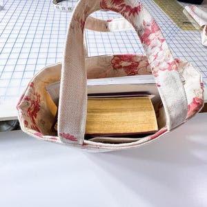 May include: A floral print tote bag with a handle, filled with books and papers. The bag's exterior features a cream-colored fabric with red floral patterns. The interior lining also has a floral design. The bag is sitting on a white surface.