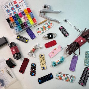Peut inclure: Une variété d'organisateurs de cordons en tissu colorés avec différents motifs et designs. Les organisateurs sont en tissu et ont une fermeture à bouton. Il y a aussi des outils pour fabriquer les organisateurs, y compris une presse à boutons et une variété de boutons.