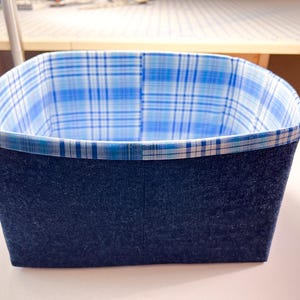 以下が含まれることがあります： 濃い青色の外側と青と白のチェック柄の内側を備えた長方形の収納バスケット。バスケットの縁は、お揃いのチェック柄の生地でトリミングされています。バスケットは、おそらくアイテムを整理するために使用されます。