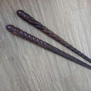 May include: Two dark brown wooden hair sticks with rounded ends. One stick features a smooth, spiralled, twisted design along its length. The second stick displays a similar twisted pattern combined with a series of small, carved leaf-like motifs.
