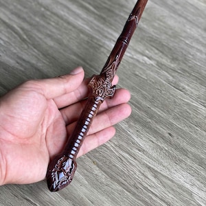 May include: A dark brown wooden wand featuring a textured, ribbed handle and an ornate, carved pommel at the base. The shaft tapers to a point, and the grip area includes intricate, raised decorative patterns near the transition to the handle.